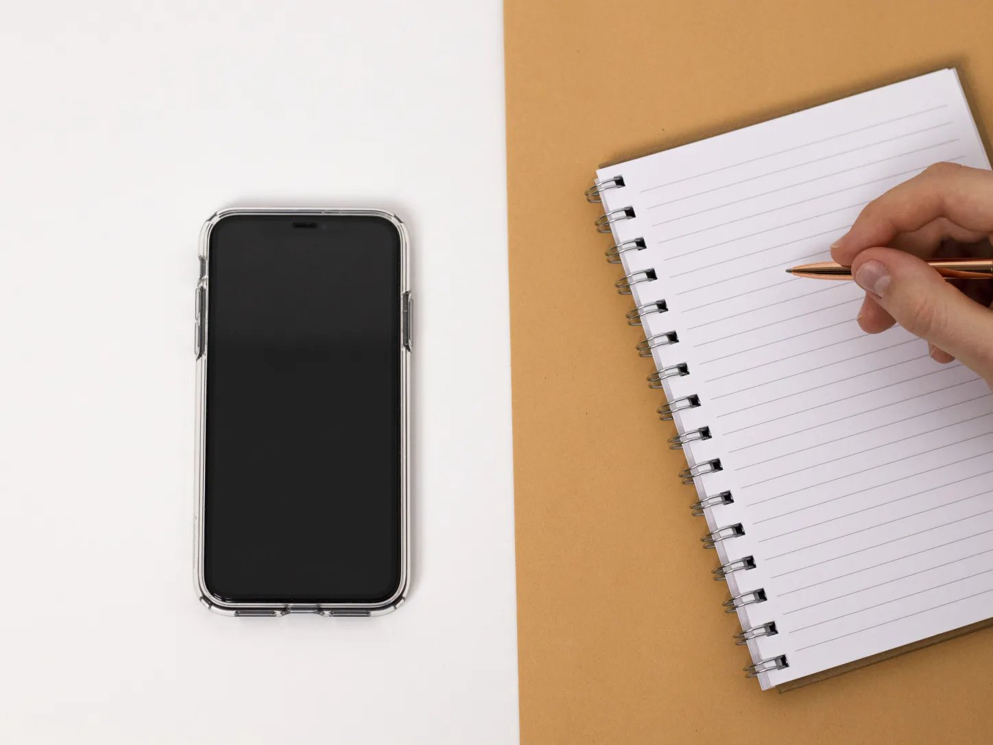 Dłoń z długopisem przy notesie i telefonie z wygaszonym ekranem